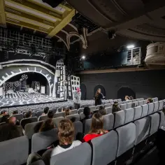 Widownia teatru z widokiem na scenę w stylu ekspresjonistycznym. Ludzie siedzą na miejscach, a reżyser omawia szczegóły z aktorką. Wnętrze foyer zaprasza do świata sztuki.
