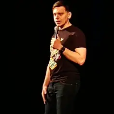 Sebastian Rejent na scenie, trzymający mikrofon. Mężczyzna w czarnej koszulce z kolorowym nadrukiem i jeansach.