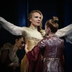 Anna Guzik w złotej sukni, otoczona przez dwie tancerki w teatralnym ujęciu.