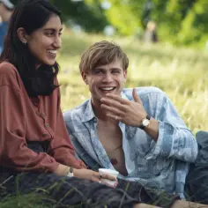 Uśmiechnięta para siedzi na trawie. To może być scena z filmu, gdzie jeden dzień obsada relaksuje się na pikniku.