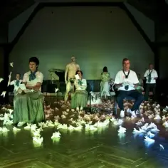 Scena teatralna pełna papierowych ptaków, ludzie w dziwnych strojach, posąg. To wszystko tworzy atmosferę teatru absurdu.