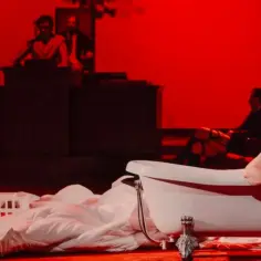 Scena teatralna z kobietą w wannie i młodą dziewczyną przy miniaturowym budynku. Intensywne czerwone oświetlenie podkreśla atmosferę performance'u.