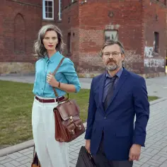 Jacek Beler i jego żona, elegancko ubrani, stoją przed ceglanym budynkiem.