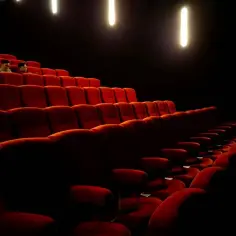 Pusta sala kinowa z rzędami czerwonych foteli. Kilka osób siedzi w oddali, tworząc intymną scenę.