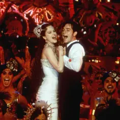 Para w białej sukni i czarnym fraku tańczy na scenie Moulin Rouge, otoczona przez tancerzy w barwnych strojach.