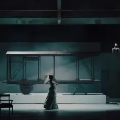 Scena teatralna z kobietą w sukni i parasolką, obok wagonu kolejowego. Nowa lalka obsada czeka na swój debiut.