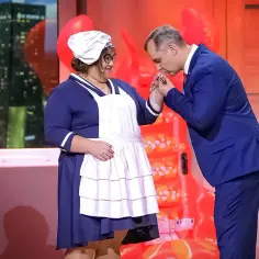 Scena jak z wodewilu: mężczyzna w garniturze całuje dłoń kobiecie w stroju pokojówki. W tle okno z widokiem na miasto i narty.