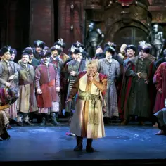 Scena z operetki: artyści w barwnych strojach historycznych, jeden klęczy, drugi gestykuluje.