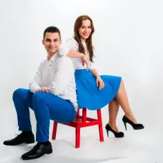 Para z kabaretu FiFa RaFa w białych koszulach i niebieskich spodniach/spódnicy siedzi na czerwonym krześle.