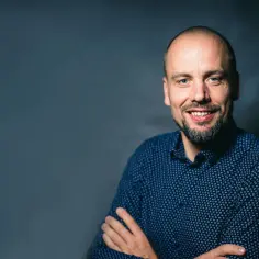 Tomasz Nowaczyk, z zarostem i w niebieskiej koszuli w drobne wzory, uśmiecha się do obiektywu, ręce skrzyżowane na piersi.