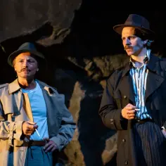 Dwóch mężczyzn w kapeluszach i prochowcach, trzymających papierosy, tworzy scenę pełną napięcia, jak z filmu melodramat.