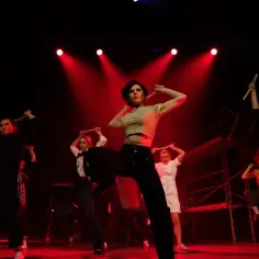 Tancerki z pałeczkami na scenie, w klimacie musicalu 1989. Czerwone światła podkreślają dynamikę sceny.