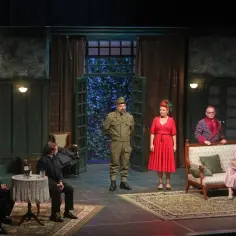Na scenie Teatru Capitol trwa dramatyczna rozmowa. Żołnierz w mundurze i kobieta w czerwonej sukni stoją pośrodku, podczas gdy inni bohaterowie siedzą lub stoją w tle, tworząc napiętą atmosferę.