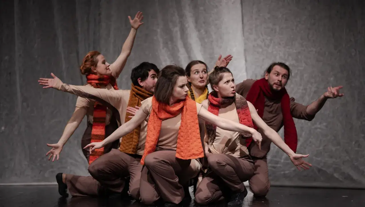 Siedmioro tancerzy w beżowych strojach i kolorowych szalikach wykonuje dynamiczną choreografię, gestykulując rękami.