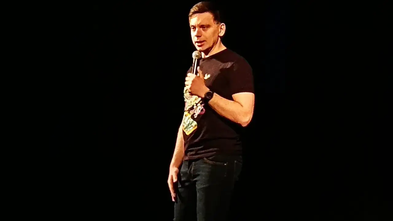 Sebastian Rejent na scenie, trzymający mikrofon. Ma na sobie czarną koszulkę z kolorowym nadrukiem i dżinsy.