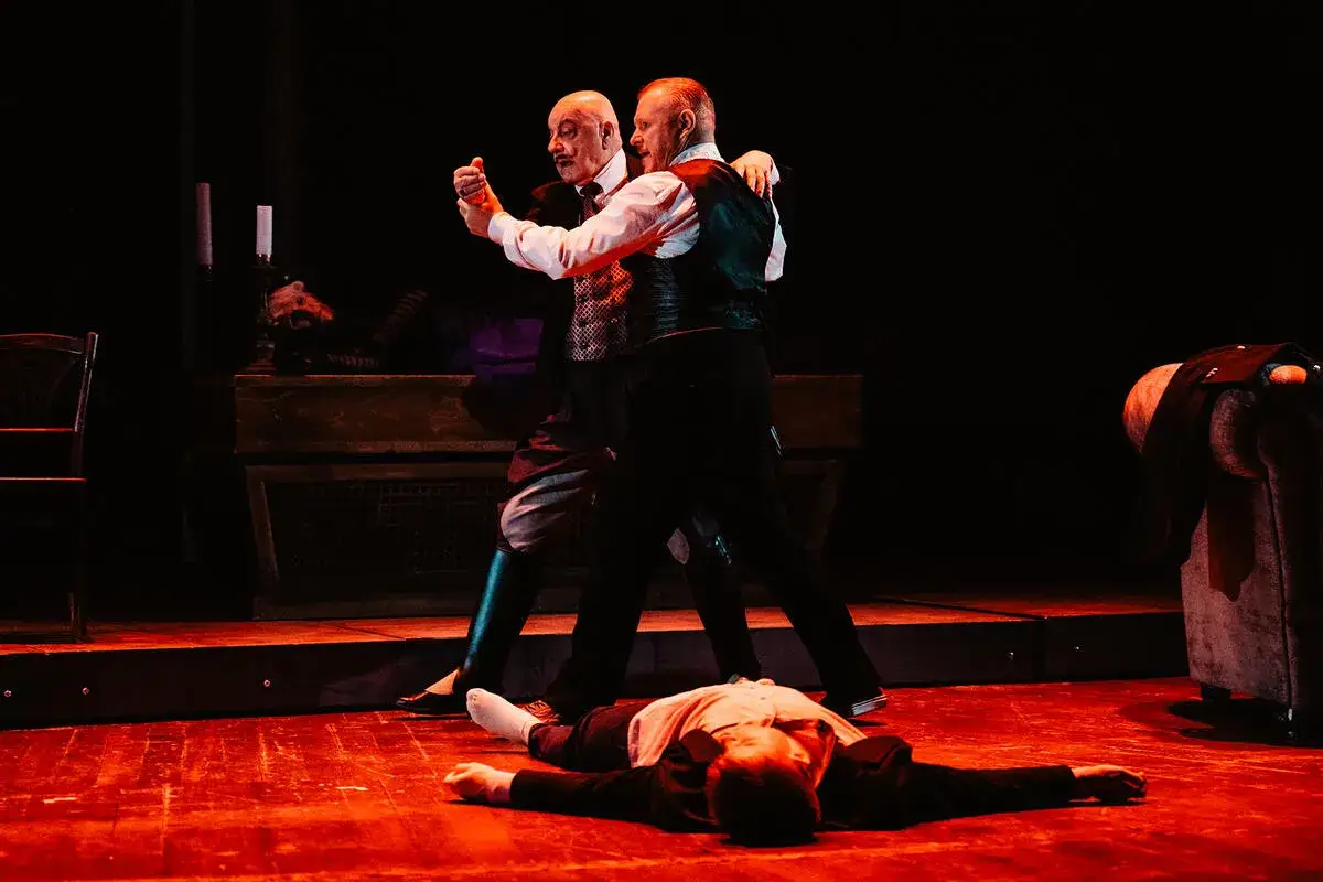 Dwóch mężczyzn tańczy tango na scenie. Na pierwszym planie leży trzeci, nieruchomy.