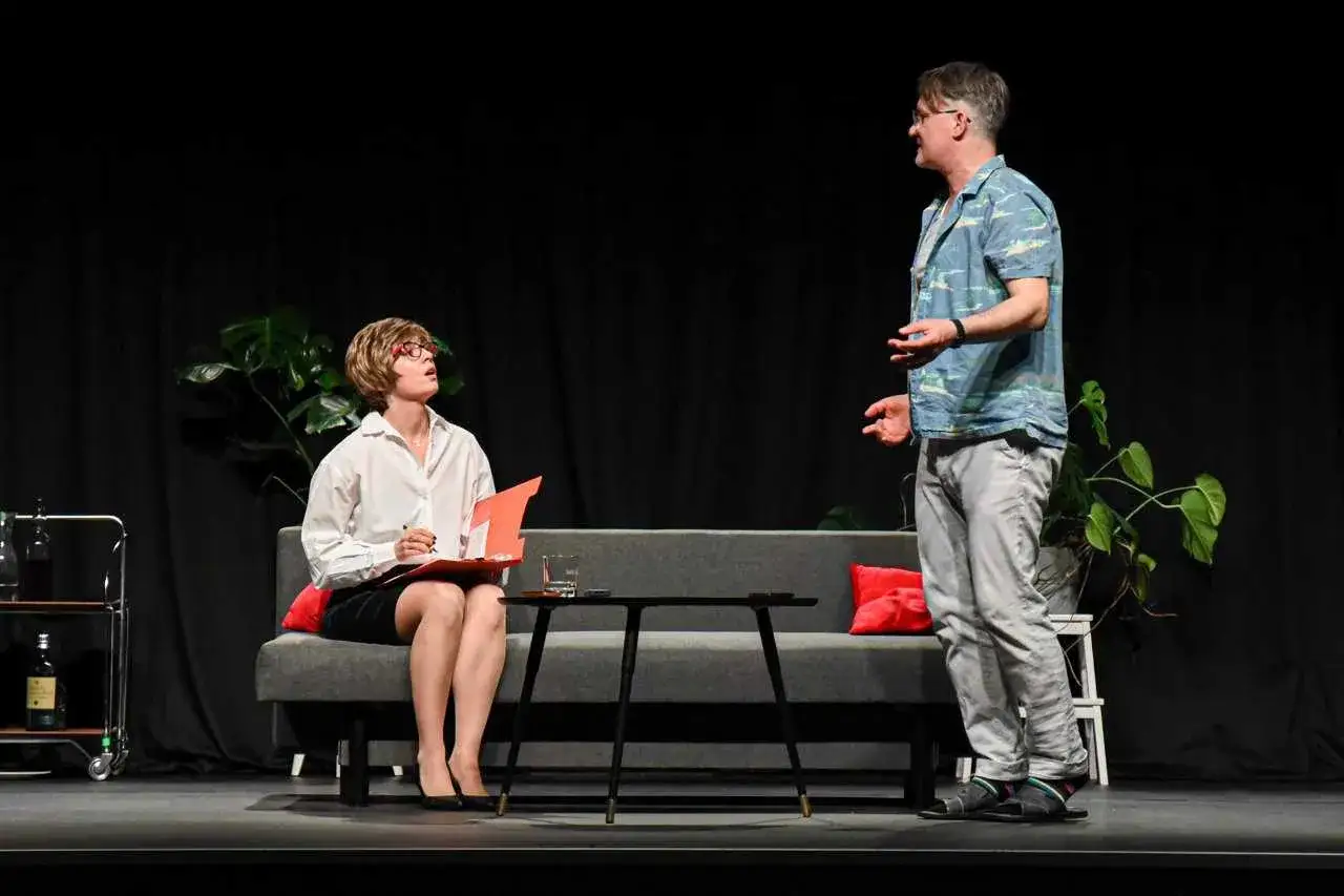 Scena teatralna: kobieta w okularach siedzi na kanapie z teczką, mężczyzna stoi obok. Czyżby rozmawiali o tym, jak znaleźć żonę do adopcji?