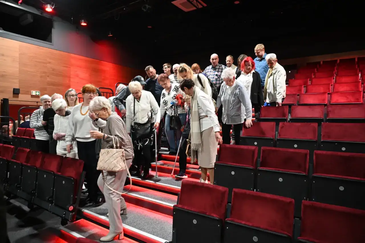 Grupa osób wchodzi po schodach w czerwonym foyer teatru, zajmując miejsca na widowni.