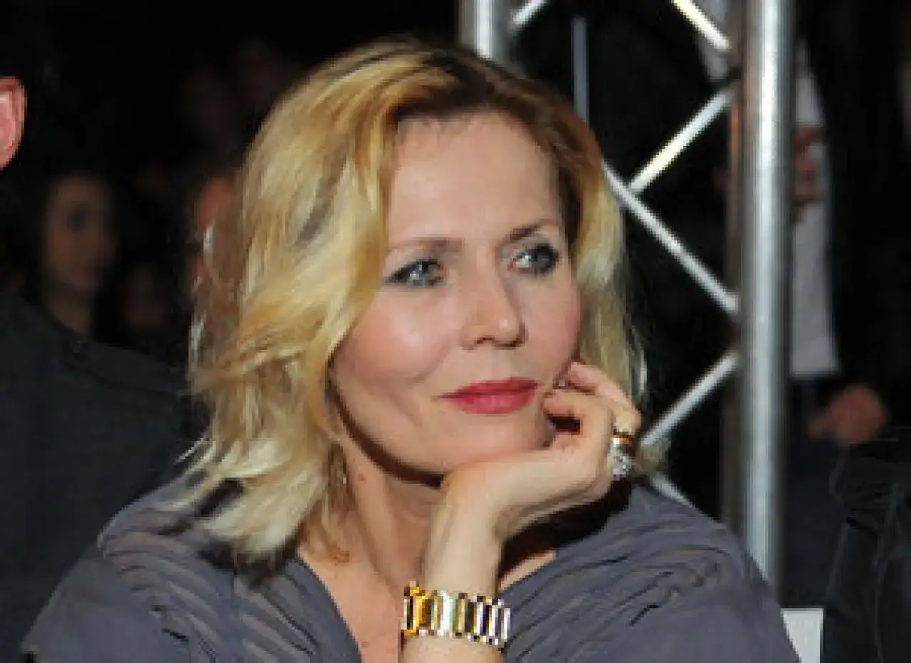 Elegancka polska aktorka po 50. z blond włosami i czerwoną szminką, opiera dłoń na policzku.