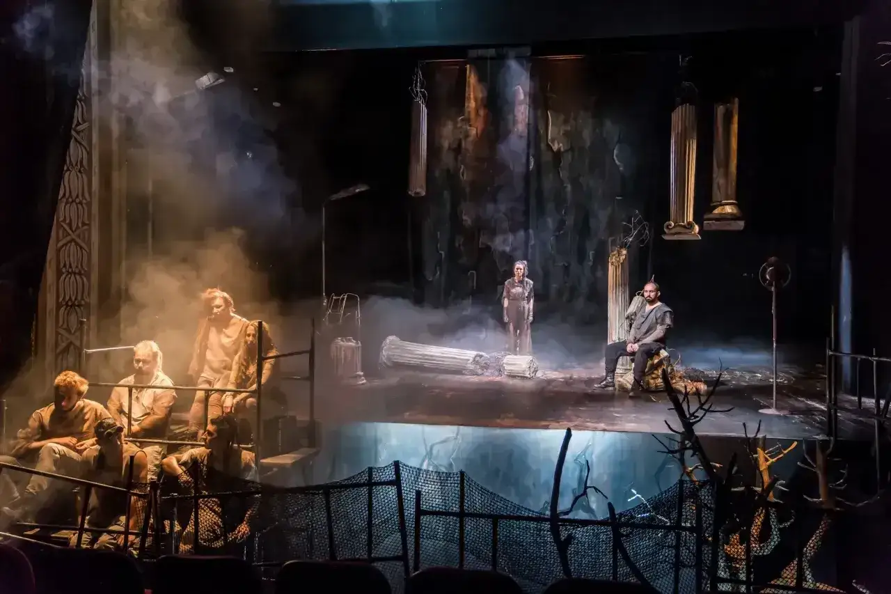 Scena teatralna z dymem i postaciami w antycznych strojach. W tle zniszczone kolumny, sugerujące konflikt tragiczny.