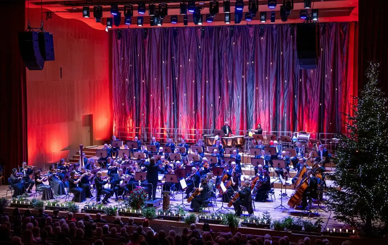 Orkiestra symfoniczna przygotowuje się do wykonania operetki. Na scenie świąteczna choinka, czerwone zasłony i oświetlenie.