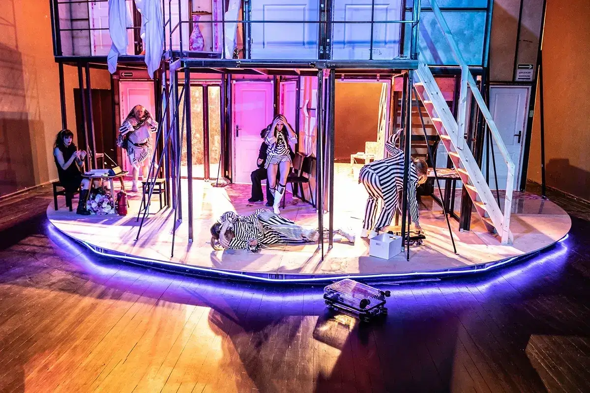 Na scenie teatralnej, w surrealistycznej scenografii, aktorzy w pasiaste stroje odgrywają scenę. O co biega Capitol?